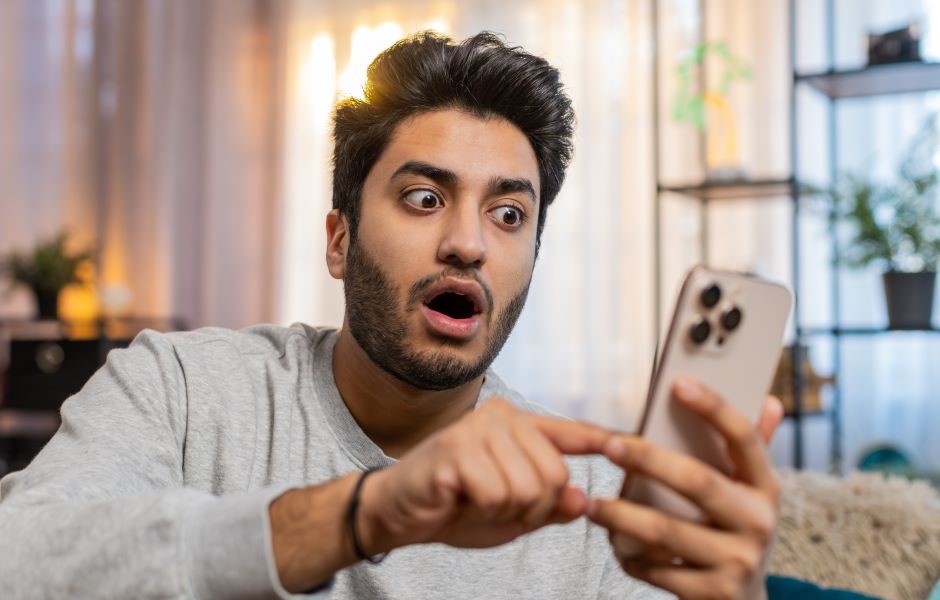 Homem surpreso usando celular em casa, ilustrando a experiência de diversão e surpresa ao usar um smartphone com tecnologia moderna.