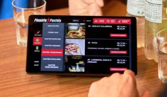 Pessoa fazendo pedido em um tablet de pizzaria, exibindo opções de pizzas como calabresa, tomate, orégano e queijo, com garrafas de água ao redor.