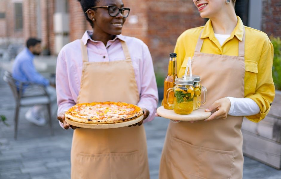 Duas mulheres sorridentes usando aventais, uma segurando uma pizza e a outra uma jarra de limonada, em ambiente ao ar livre, promovendo comida caseira.