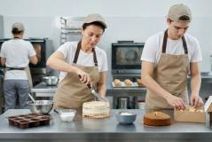 Três confeiteiros de camiseta branca, avental bege e boné trabalham em uma cozinha industrial. No primeiro plano, uma mulher espalha recheio em um bolo com espátula enquanto um rapaz embala doces em uma caixa de papelão. Ao fundo, uma terceira pessoa trabalha de costas perto de fornos e prateleiras de inox.