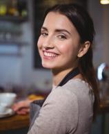 Mulher jovem sorridente, usando avental, em frente ao balcão de uma cafeteria. A imagem transmite hospitalidade, bom atendimento e profissionalismo no setor de alimentação.
