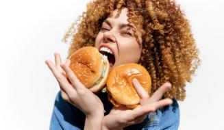 Mulher sorridente com cabelo cacheado dourado segurando dois hambúrgueres e expressando alegria,