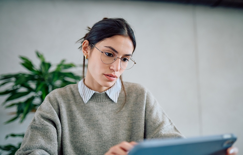 Mulher estudando com tablet em ambiente moderno, usando óculos e blusa cinza, com expressão concentrada e fundo de parede clara e planta verde ao fundo.