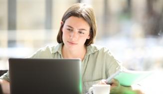 Uma jovem mulher concentrada estudando no computador em um café, com caderno e caneta ao lado, refletindo foco e produtividade. Ambiente iluminado e aconchegante.