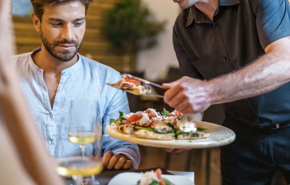 Um homem em um restaurante recebe um prato com comida de um garçom, enquanto uma mulher senta ao lado, com um copo de bebida.