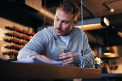 Homem concentrado escrevendo em um restaurante contemporâneo, usando uma roupa casual e observando atentamente documentos ou menu, ambiente acolhedor com iluminação quente.