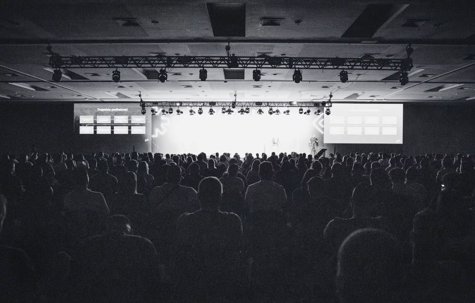 Grande plateia assistindo a uma apresentação ou palestra em um evento corporativo ou conferência, com iluminação focada no palco e telas de projeção ao fundo.