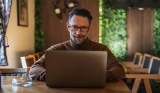 Homem de meia idade com barba, usando óculos, trabalhando em um laptop em um ambiente acolhedor e bem iluminado, com móveis de madeira e plantas ao fundo.