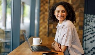 Mulher sorridente com cabelo cacheado no ambiente de um escritório, usando camisa listrada, ao lado de um laptop, uma xícara de café e um celular, representando profissionalismo e produtividade.