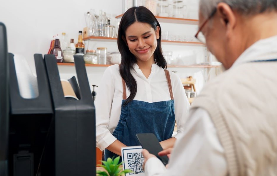Jovem mulher sorridente fazendo pagamento com smartphone em uma cafeteria, usando uniforme e atendente ao lado, ambiente acolhedor e bem decorado