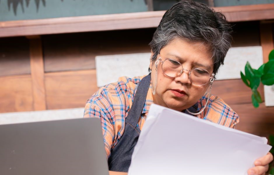 Mulher com óculos lendo um documento de forma concentrada, em ambiente de escritório com decoração de madeira e plantas ao fundo.