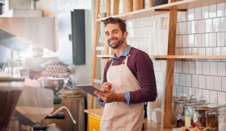 Homem sorridente com barba, vestindo avental e camisa azul, trabalhando em uma cafeteria ou café, segurando um tablet com prateleiras de produtos ao fundo.