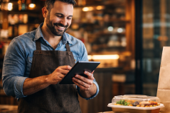 Empreendedor de delivery usando tablet para gerenciar pedidos e atualizar o cardápio, com embalagem de comida pronta para entrega ao lado.