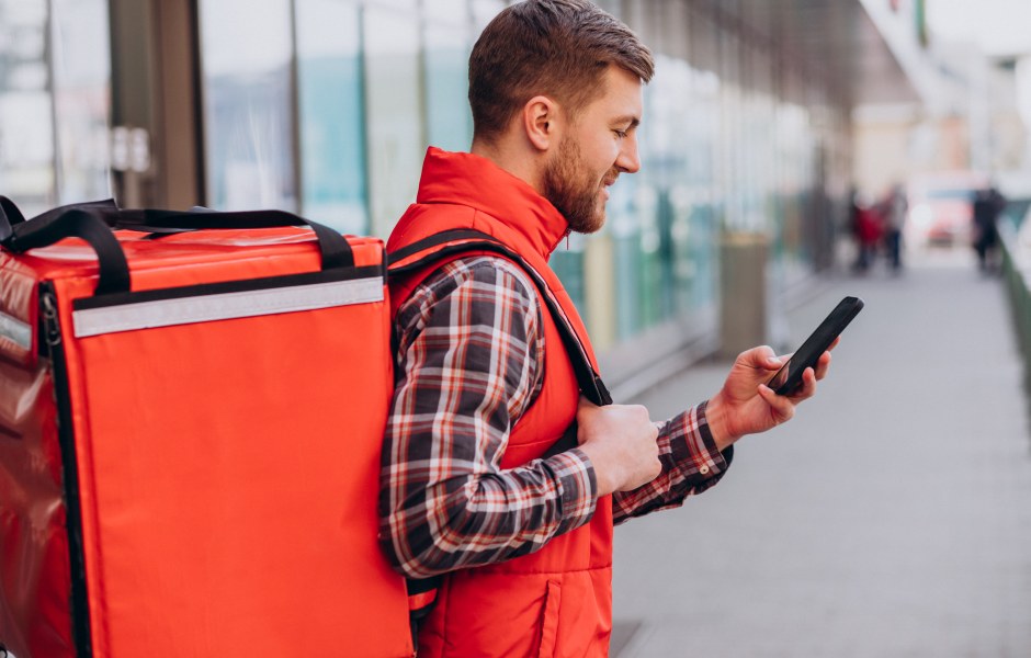Entregador de colete vermelho e camisa xadrez, visto de perfil em uma calçada, carrega uma grande mochila de delivery nas costas enquanto mexe em seu smartphone.