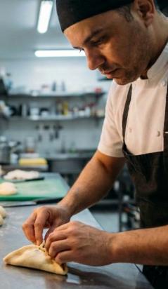 Um cozinheiro de avental e bandana preta trabalha concentrado na bancada de inox de uma cozinha profissional, moldando manualmente uma massa recheada (estilo esfiha ou pastel), cercado por bowls com ingredientes e porções de massa prontas.