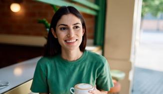 Mulher sorridente com cabelo castanho escuro, vestindo uma camiseta verde, segurando uma xícara de café ou chá, sentado em um café ou restaurante bem iluminado, com ambiente acolhedor e decorado com elementos naturais.
