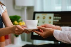 A mulher recebendo uma xícara de café de um atendente em um ambiente interno, com computador ao fundo, simbolizando serviço de cafeteria ou entrega de bebidas