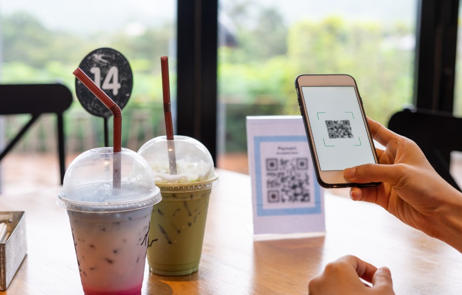 Imagem de uma pessoa usando um smartphone para escanear um código QR em um café, com duas bebidas em destaque na mesa.