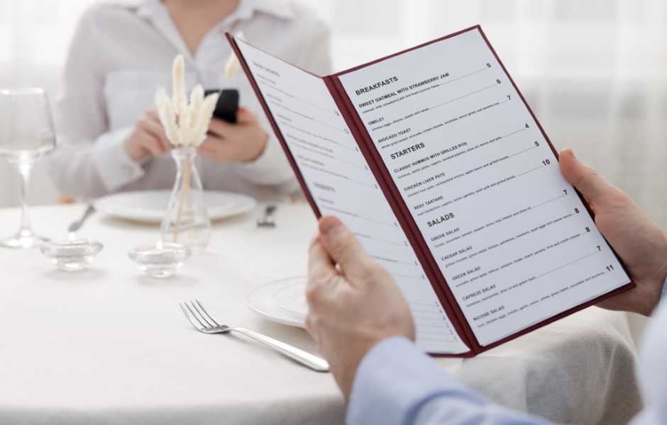 Um homem olhando o cardápio em um restaurante enquanto uma mulher usa o celular ao fundo, com uma mesa elegantemente arrumada.