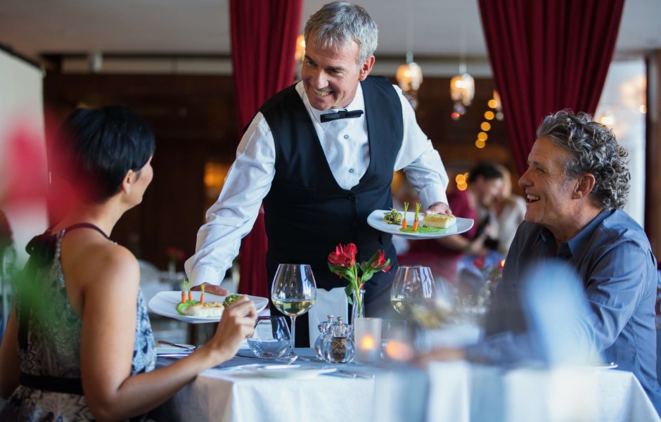 Um garçom serve pratos para um casal em um restaurante elegante, com flores e vinho na mesa, criando um ambiente romântico.