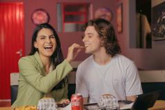 Casal sorridente compartilhando um momento divertido em uma lanchonete, saboreando comida e bebida, com um ambiente descontraído ao fundo.