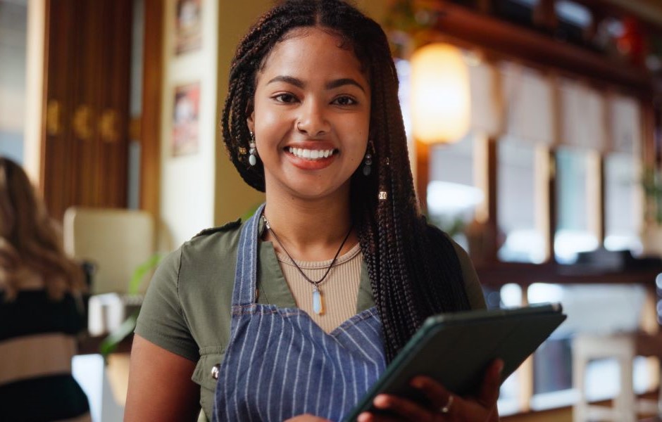 Mulher sorridente com cabelo trançado, usando avental no ambiente de um restaurante ou café, segurando um tablet, no contexto de trabalho na área de gastronomia ou atendimento ao cliente.