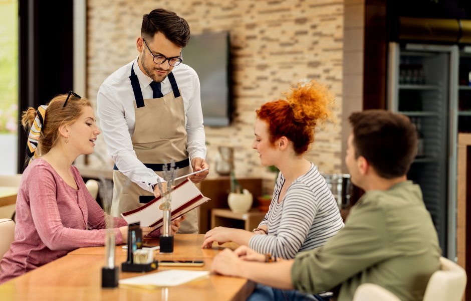 Garçom de avental bege e óculos atende três clientes sentados à mesa de um restaurante, apontando para as opções em um cardápio aberto.