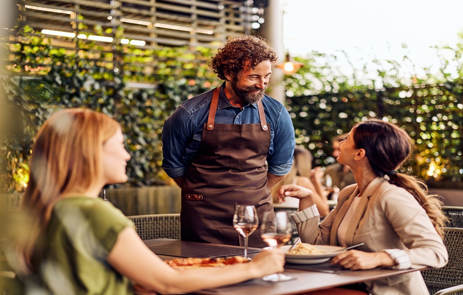 Homem com barba e cabelo cacheado atendendo duas mulheres em um restaurante ao ar livre, com pratos de comida e taças de vinho, ambiente acolhedor e natural.
