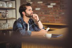 Homem jovem de barba e camisa xadrez azul, sentado em uma mesa de café. Ele apoia a mão no queixo e olha para o lado com expressão séria. À sua frente estão um celular na mão, uma xícara de café e um tablet.