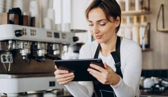 Barista sorridente de avental preto opera um tablet com as duas mãos em um balcão de cafeteria. Ao fundo, destaca-se uma máquina de café expresso profissional branca.