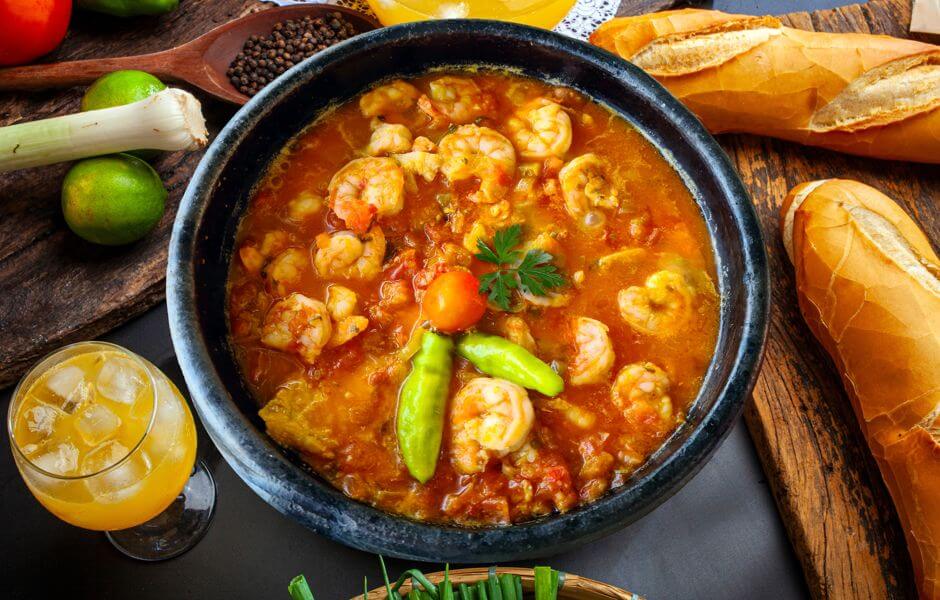 Uma deliciosa moqueca de camarão em um prato escuro, acompanhada de pães e uma bebida refrescante.