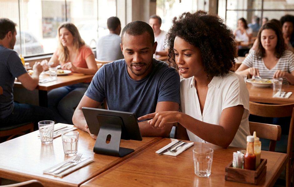 Casal está sentado em uma mesa de restaurante movimentado interagindo com um tablet fixado em um suporte. A mulher negra, de cabelo cacheado e camisa branca, aponta para a tela enquanto o homem ao seu lado observa atento. Sobre a mesa de madeira, há copos de água, talheres dispostos em guardanapos e um suporte com molhos.