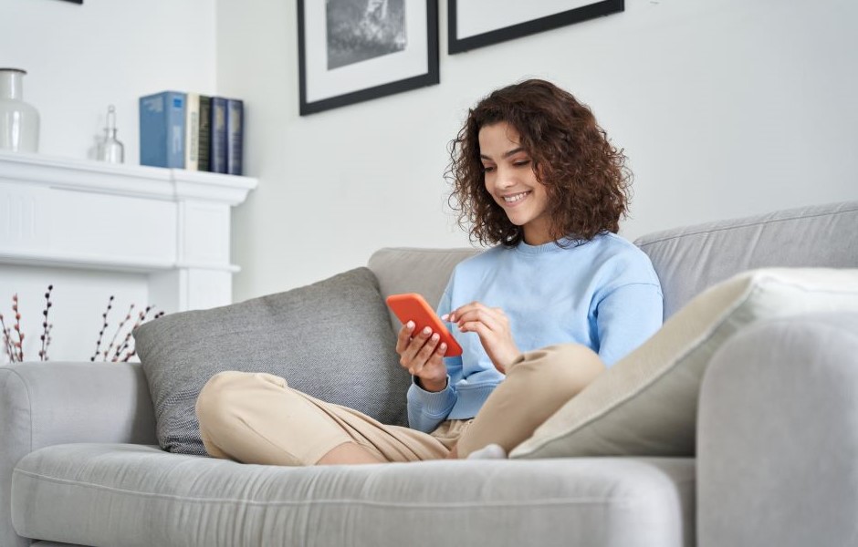 Jovem sorridente usando celular enquanto relaxa no sofá de casa, ambiente moderno e aconchegante, ideal para o conforto e entretenimento.