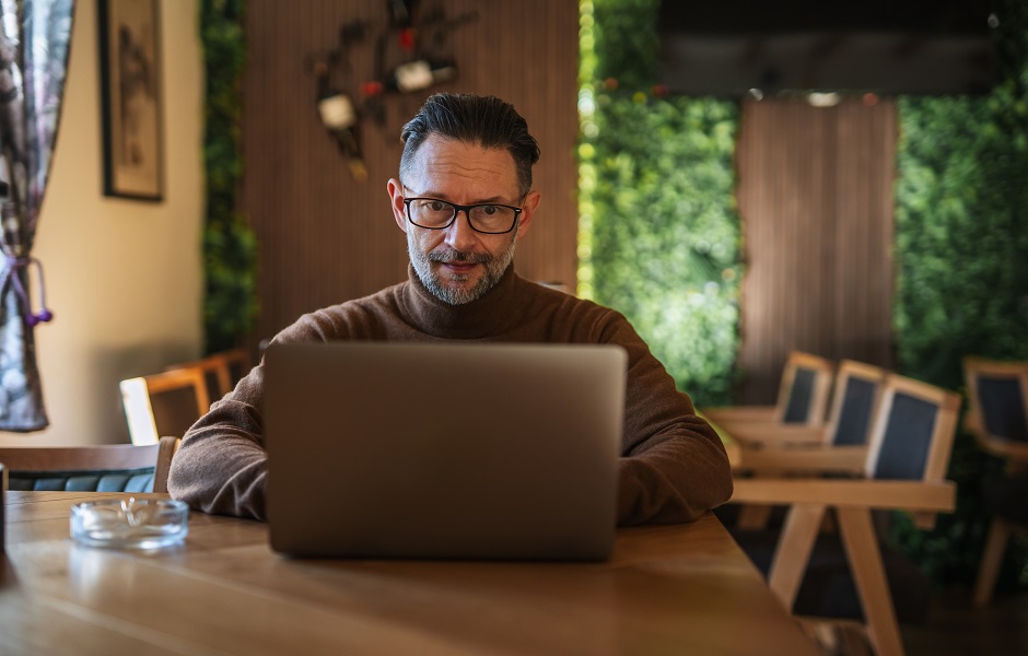 Homem de meia idade com barba, usando óculos, trabalhando em um laptop em um ambiente acolhedor e bem iluminado, com móveis de madeira e plantas ao fundo.