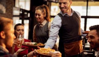 Garçom e garçonete sorrindo servindo pratos de comida e bebida a clientes em um restaurante com ambiente aconchegante.