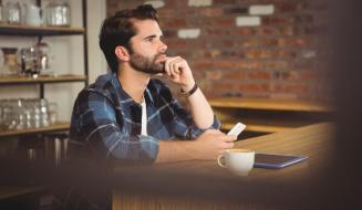 Homem jovem de barba e camisa xadrez azul, sentado em uma mesa de café. Ele apoia a mão no queixo e olha para o lado com expressão séria. À sua frente estão um celular na mão, uma xícara de café e um tablet.