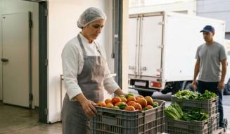 Mulher com avental e touca segura uma caixa plástica com tomates e pimentões em um entreposto ou cozinha comercial, com um homem e um caminhão de entregas ao fundo.