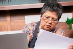 Mulher com óculos lendo um documento de forma concentrada, em ambiente de escritório com decoração de madeira e plantas ao fundo.