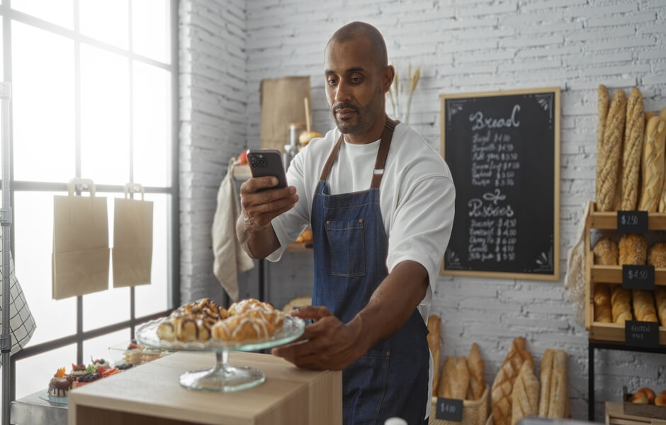 Homem em uma padaria verificando seu celular enquanto serve doces em um prato. Ambiente acolhedor com pães e quitutes ao fundo.