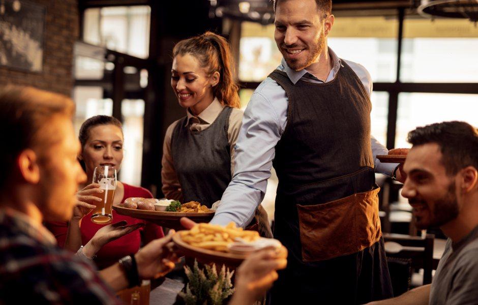Garçom e garçonete sorrindo servindo pratos de comida e bebida a clientes em um restaurante com ambiente aconchegante.