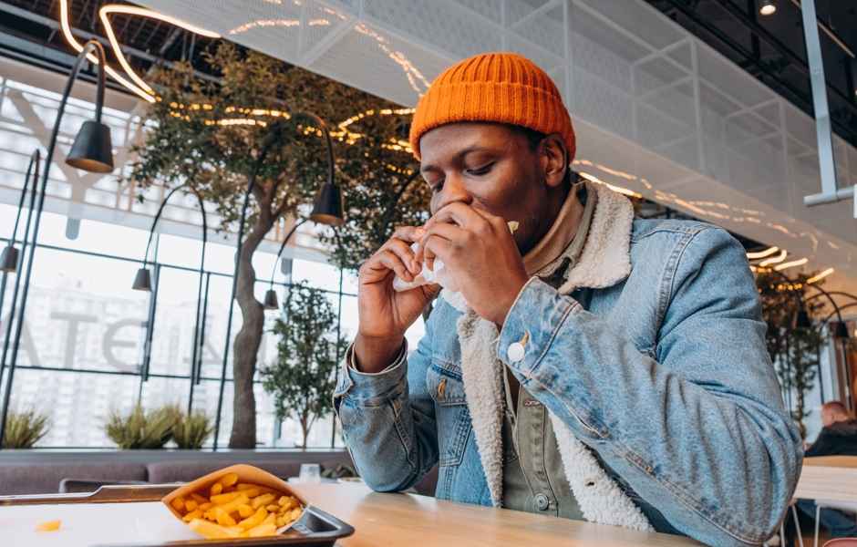 Homem com boné laranja saboreando um sanduíche em um café moderno, cercado por plantas e decoração contemporânea, com batatas fritas ao lado.