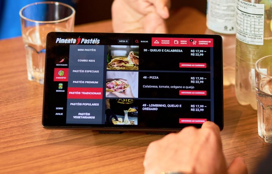 Pessoa fazendo pedido em um tablet de pizzaria, exibindo opções de pizzas como calabresa, tomate, orégano e queijo, com garrafas de água ao redor.