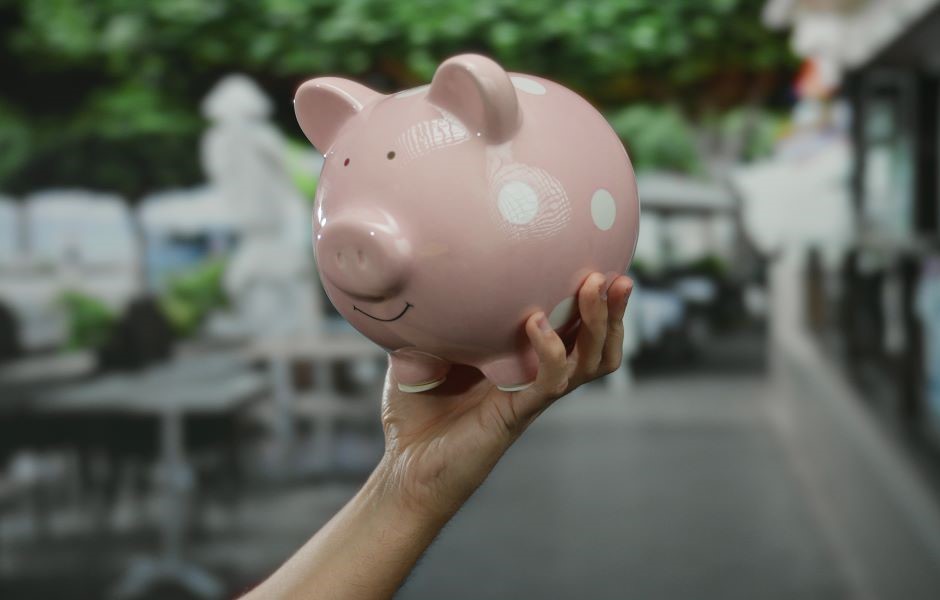 Pessoa segurando uma vaquinha de porcelana rosa com detalhes brancos em um ambiente ao ar livre, simbolizando economia doméstica ou poupança.