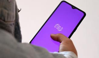 Imagem de um smartphone exibindo o aplicativo Nubank com fundo roxo, mostrando a logo e a interface digital. O usuário interage com a tela, destacando a modernidade e praticidade do aplicativo de serviços financeiros.
