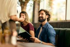 Homem sorridente em um restaurante ou café, interagindo com um garçom enquanto lê um cardápio, ao lado de uma mulher. Ambiente aconchegante com grande janela.