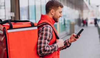 Entregador de colete vermelho e camisa xadrez, visto de perfil em uma calçada, carrega uma grande mochila de delivery nas costas enquanto mexe em seu smartphone.