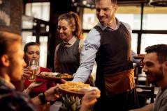 Garçom e garçonete sorrindo servindo pratos de comida e bebida a clientes em um restaurante com ambiente aconchegante.