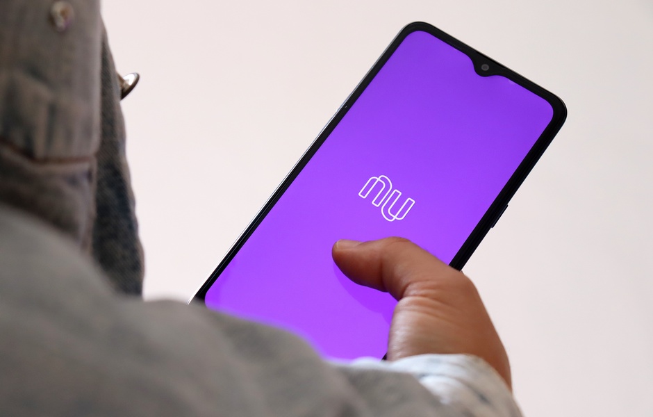Imagem de um smartphone exibindo o aplicativo Nubank com fundo roxo, mostrando a logo e a interface digital. O usuário interage com a tela, destacando a modernidade e praticidade do aplicativo de serviços financeiros.