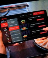 Pessoa usando um tablet para fazer pedido em um restaurante de comida japonesa, com pratos de sashimi e salmão visíveis na mesa, destacando o uso de tecnologia na experiência gastronômica.
