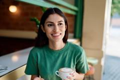 Mulher sorridente com cabelo castanho escuro, vestindo uma camiseta verde, segurando uma xícara de café ou chá, sentado em um café ou restaurante bem iluminado, com ambiente acolhedor e decorado com elementos naturais.